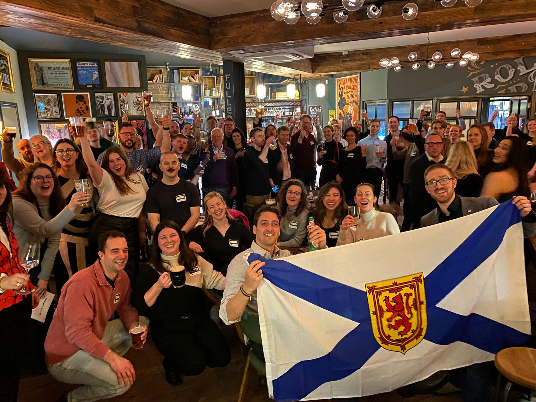 A large group of people in a pub, looking at the camera smiling while raising their glasses. In the foreground, a large Nova Scotia flag is being held up.