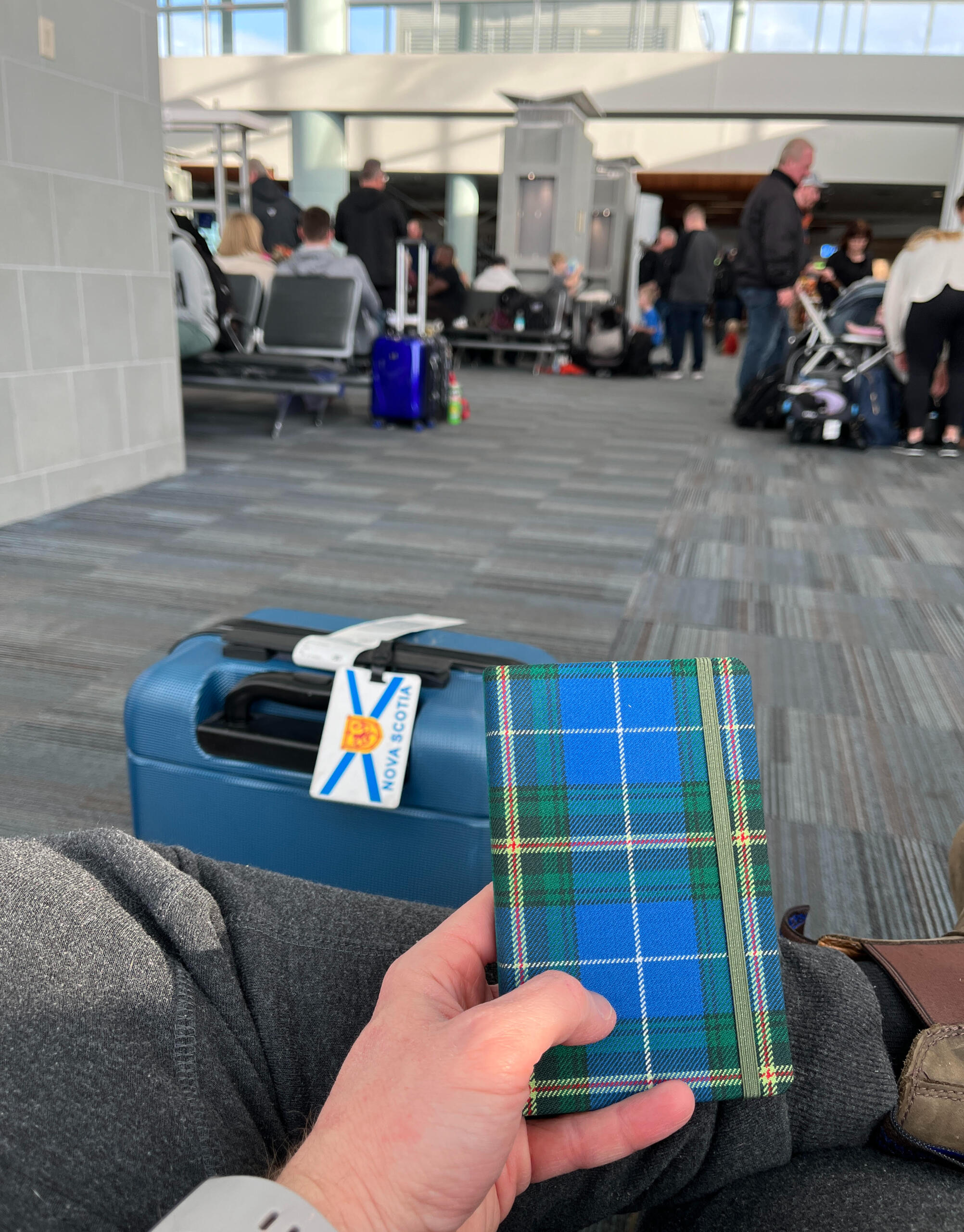 Photo is a first person perspective. The photographer is sitting in an airport and in the foreground of the photo, they are holding a small notebook that is patterned in the Nova Scotia tartan. In the mid-ground, is a piece of luggage with a Nova Scotia fl